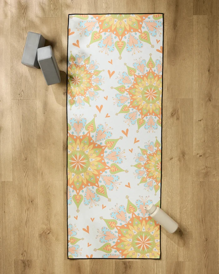 The Mindful Mandalas yoga mat by Mindfulnice features vibrant orange, yellow, and green colors on a wooden floor, paired with two gray yoga blocks and a white water bottle. A non-slip towel enhances grip and comfort for your practice.