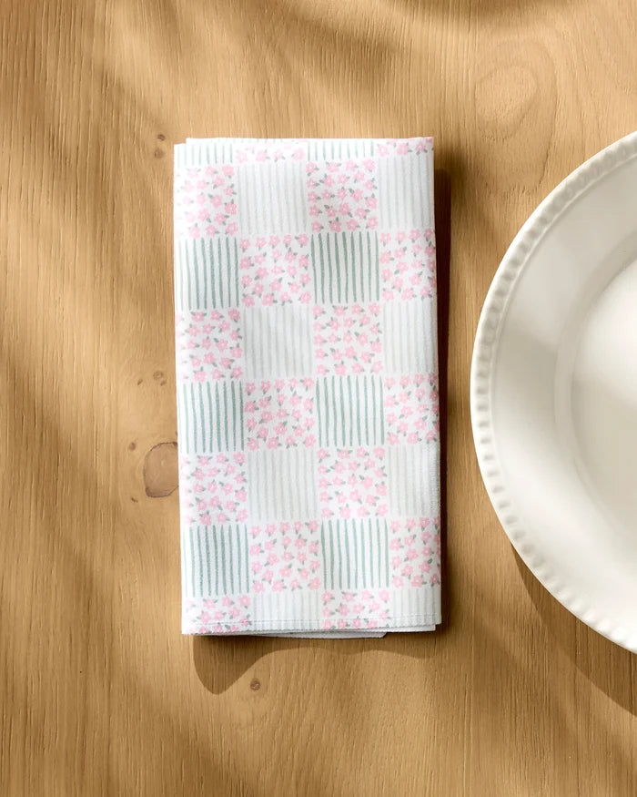 A Hufton Studio Mini Flowers and Stripes Check folded cloth napkin, featuring a pink floral and light blue striped checkerboard pattern, sits on a wooden table beside a scalloped white plate.