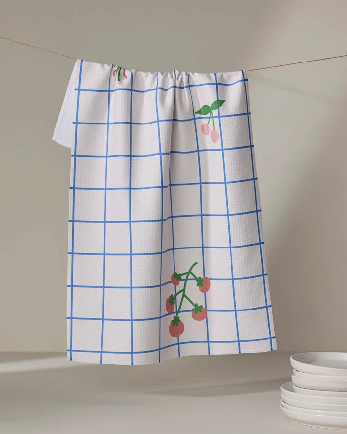 The Mini Harvest dish towel by Letters By Shells, featuring a blue grid and cherry print, hangs on a clothesline above white plates, adding charm and function to your kitchen.