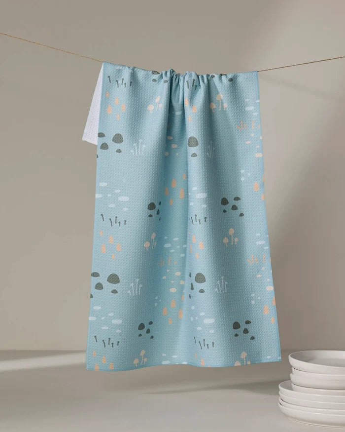 The Amberleigh Adoff Mini Shroom Sprouts kitchen towel, crafted from recycled materials, displays a pastel mushroom and plant pattern in light blue and hangs above white plates against a neutral background.