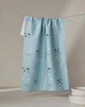 The Amberleigh Adoff Mini Shroom Sprouts kitchen towel, crafted from recycled materials, displays a pastel mushroom and plant pattern in light blue and hangs above white plates against a neutral background.