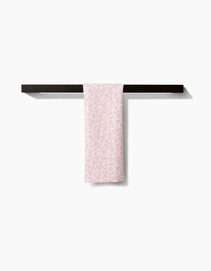 The Teri the Butter Minimalist Blush hand towel, featuring a pink botanical design and made from ultra-absorbent Geoweave fabric, hangs neatly on a dark horizontal bar against a plain white background.