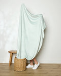 Only the lower legs and white sneakers of a person wrapped in GEOMETRY's Mint Drift throw blanket are visible as they stand on a woven basket. A wooden stool sits nearby, with a white wall and wood floor setting the minimalist scene.