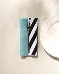 A fork rests on a GEOMETRY Mint Stripes recycled dinner napkin with teal and black-and-white diagonal stripes, placed beside a white plate on a textured light surface.
