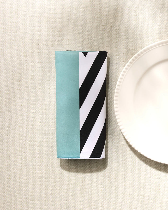 The GEOMETRY "Mint Stripes" reusable cloth napkin, featuring teal, white, and black diagonal stripes, rests neatly folded on a textured cream tablecloth beside the edge of a scalloped white plate.