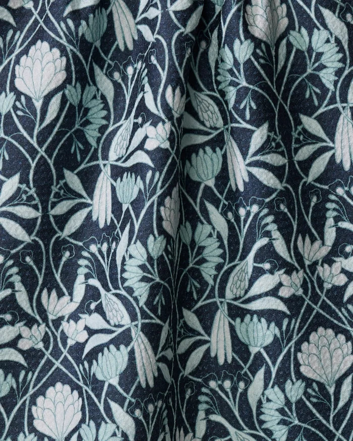 The Mirielle kitchen towel by Amy MacCready features dark fabric with a symmetrical design of light blue and pale green flowers, leaves, and birds. Made from recycled materials, it has a slightly wrinkled, textured botanical surface.