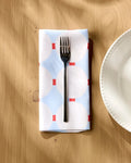 A silver fork rests on Happy Hemera’s Mist Blue Oasis dinner napkin, adorned with white circles and red lines, placed on a wooden table beside a scalloped white plate.