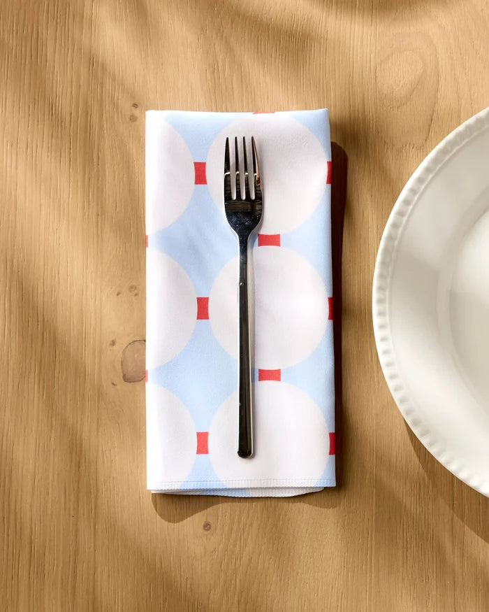 A silver fork rests on Happy Hemera’s Mist Blue Oasis dinner napkin, adorned with white circles and red lines, placed on a wooden table beside a scalloped white plate.