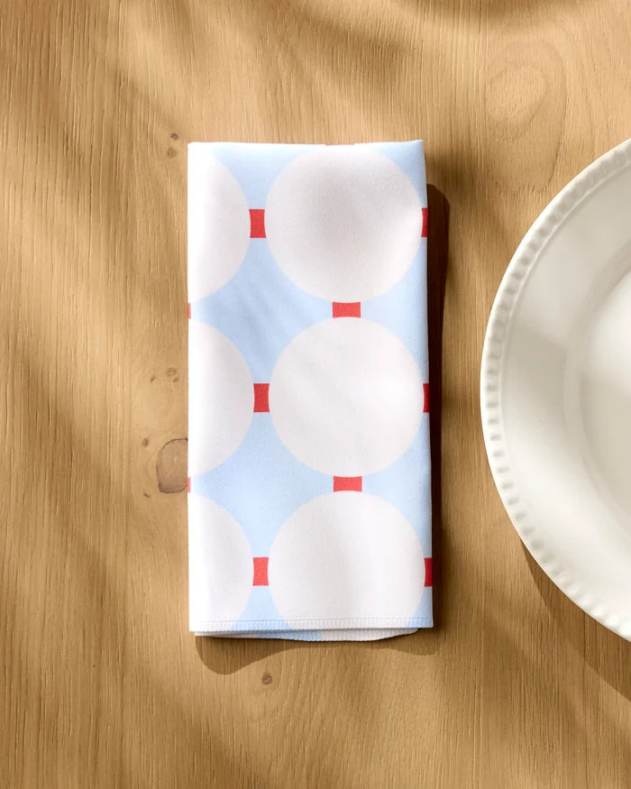 The Mist Blue Oasis reusable dinner napkin by Happy Hemera, featuring a light blue and white geometric pattern with red squares, is placed on a wooden table next to a white ceramic plate.
