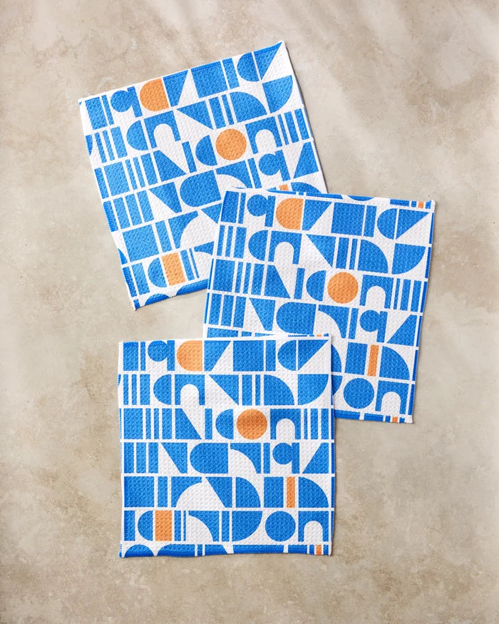 Three square Misty Oasis cloths by Happy Hemera, featuring blue geometric and orange circle patterns, are arranged on a beige marble surface. This super-absorbent dishtowel set offers a modern, abstract design with repeating shapes.