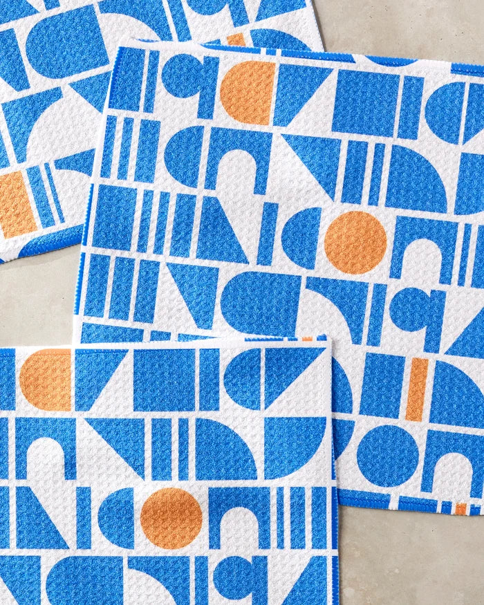 Four Misty Oasis dishtowels by Happy Hemera, featuring bold blue and orange geometric patterns—circles, semi-circles, and rectangles—are laid out on a beige surface, overlapping at the corners.