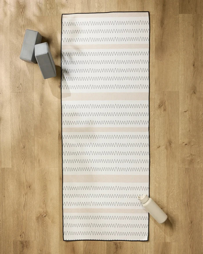 The Mod Zig Zag yoga towel by Coit Creative, featuring white non-slip fabric and zigzag patterns, is laid on a wooden floor with two gray yoga blocks in the top left corner and a white water bottle at the bottom right.