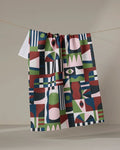 The "Modern Bauhaus Mood" recycled kitchen towel by Claire Iglesias, featuring bold green, blue, red, white, and pink geometric patterns, hangs on a clothesline against a plain beige background.