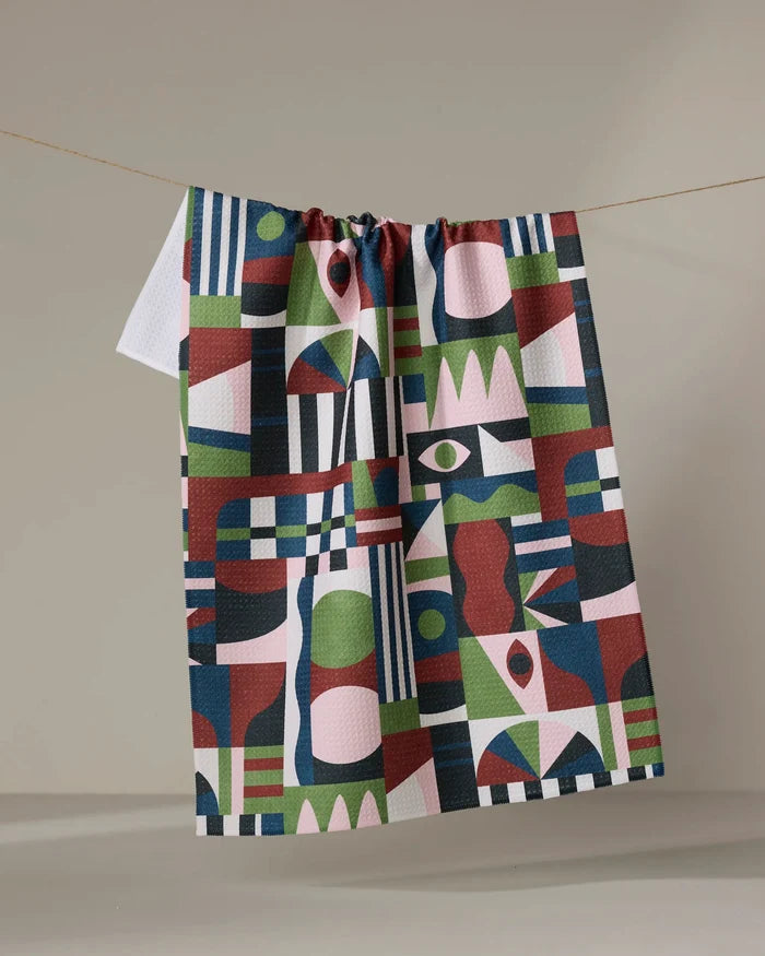 The "Modern Bauhaus Mood" recycled kitchen towel by Claire Iglesias, featuring bold green, blue, red, white, and pink geometric patterns, hangs on a clothesline against a plain beige background.