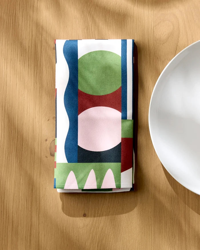 The Modern Bauhaus Mood napkin by Claire Iglesias, featuring a colorful geometric pattern in green, pink, red, and blue, is folded on a light wooden table beside a white plate.