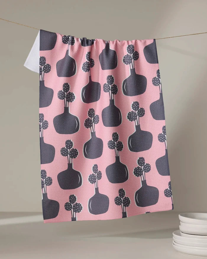 The "Modern Bouquet" tea towel by Lizzie Clark features a pink quick-drying design with dark vases and floral motifs. It hangs on a line with a folded white corner, while white plates are stacked nearby.