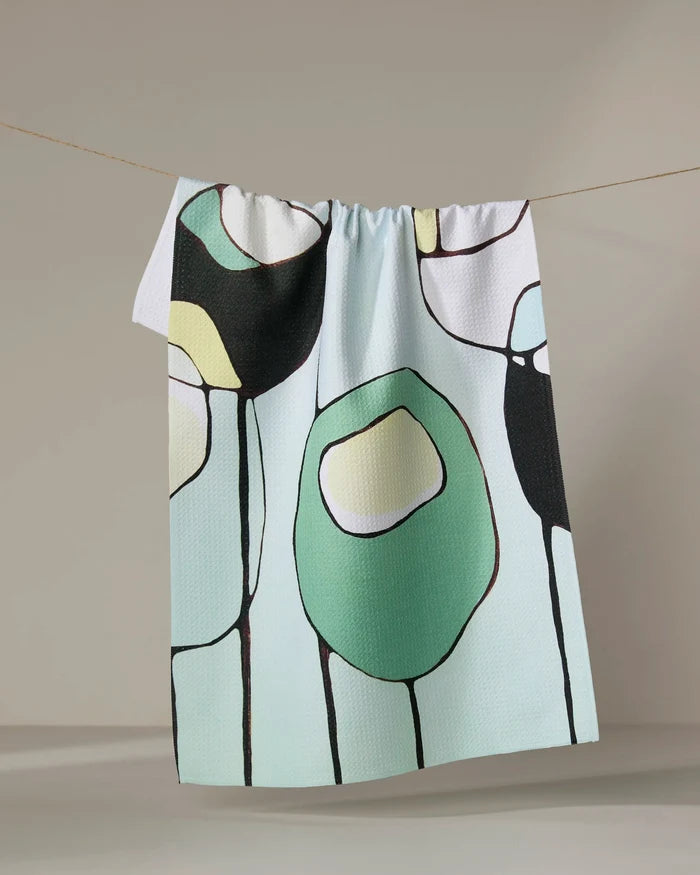 The "Modern Poppy" tea towel by Rachel Austin, featuring abstract green, black, and yellow shapes outlined in black, dries quickly and hangs on a clothesline against a neutral background.