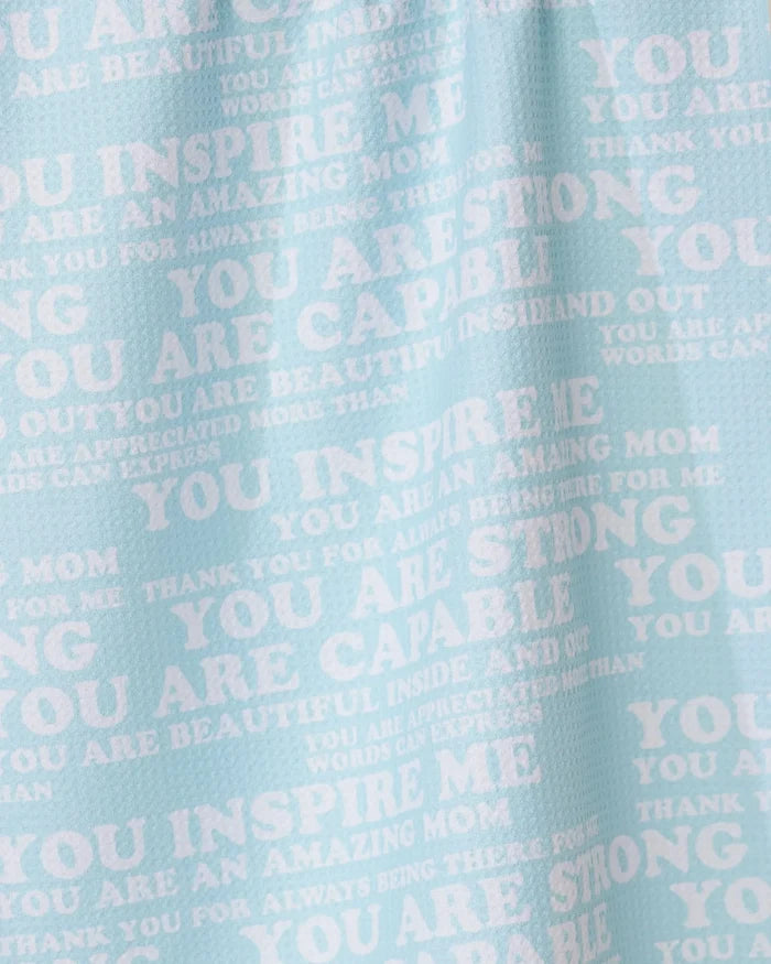 The GEOMETRY Mom Mantra tea towel is light blue, made from recycled materials, and features white overlapping motivational phrases like YOU INSPIRE ME and YOU ARE AMAZING MOM in different fonts and sizes.