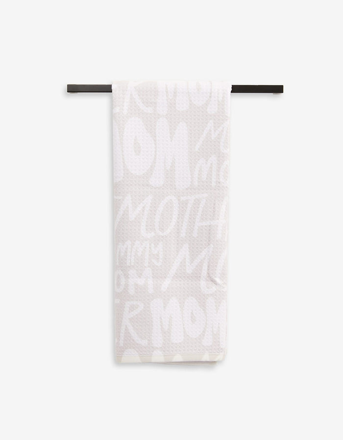 A light-colored, ultra-absorbent GEOMETRY Mom tea towel hangs on a black rod, showcasing playful white lettering with words like MOM, MOTHER, and MOMMY.