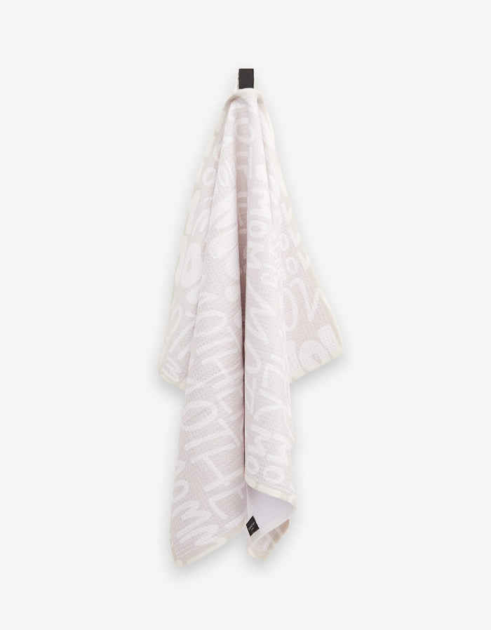 A light-colored, ultra-absorbent GEOMETRY tea towel, called “Mom,” featuring bold white abstract letters and numbers, hangs from a black hook against a plain background.