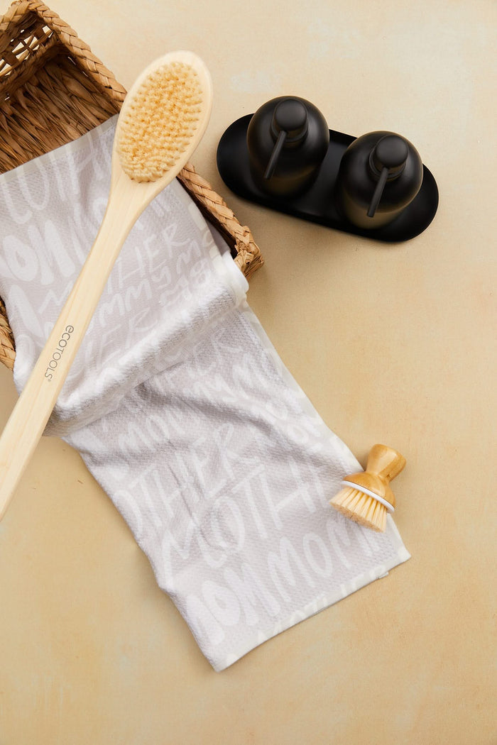A GEOMETRY Mom wooden bath brush, small scrub brush, white ultra-absorbent tea towel, and black soap dispensers sit on a beige surface; the tea towel is partially draped over a woven basket.