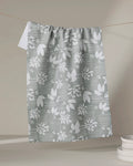 The Toboggan Avenue "Moments" kitchen tea towel, featuring a gray and white floral and leaf pattern made from recycled materials, hangs on a clothesline near a stack of white plates against a neutral background.