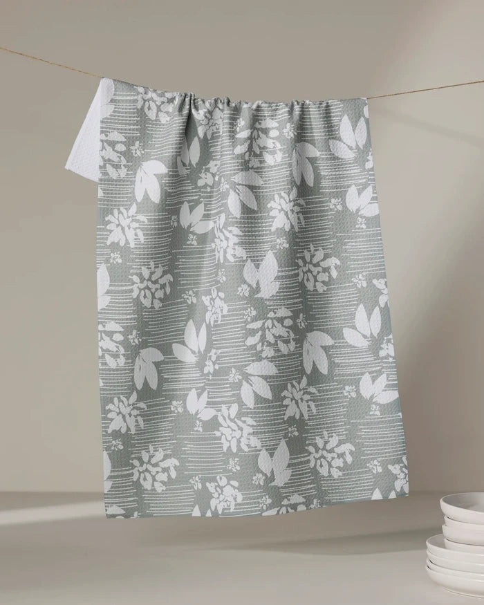 The Toboggan Avenue "Moments" kitchen tea towel, featuring a gray and white floral and leaf pattern made from recycled materials, hangs on a clothesline near a stack of white plates against a neutral background.