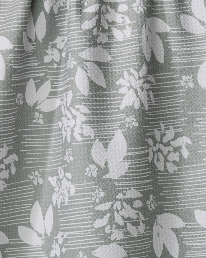 Close-up of the Moments kitchen tea towel by Toboggan Avenue, crafted from recycled materials with a white leaf and flower pattern on light gray, featuring horizontal lines and gentle folds.