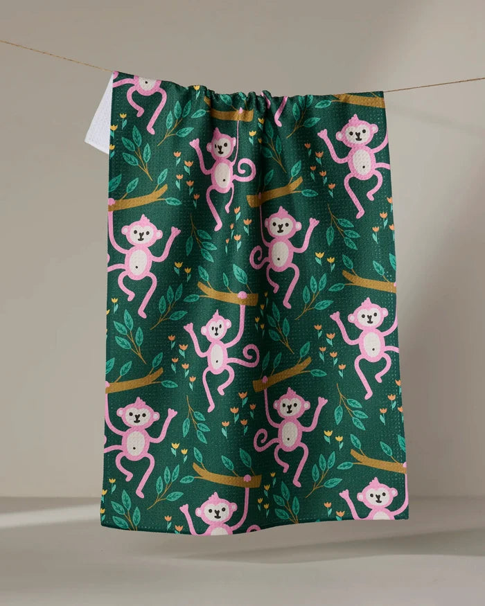 The "Monkeys" tea towel by Genna Blackburn, made from recycled materials, displays playful pink monkeys swinging among branches and leaves on a dark green background.