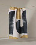 The Geometry Mono towel, featuring an abstract black and gold geometric design, hangs from a string against a neutral background.