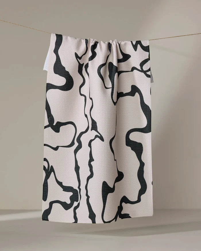 The Monochrome Liquid kitchen towel by Marina Solodka, cream-colored and recycled with abstract black squiggly lines, hangs from a string against a neutral background.