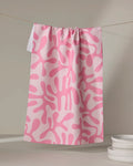 The "Monochrome Morning" tea towel by Marina Solodka, featuring an abstract pink and white splatter pattern, hangs on a clothesline. Made from recycled materials, it dries quickly. White plates are stacked in the lower right corner on a neutral background.