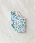 A stack of Moody Flowers napkins by Masha Volnova, made from eco-friendly paper and featuring swirling blue, pink, and green abstract foliage patterns, is arranged neatly on a marble surface.