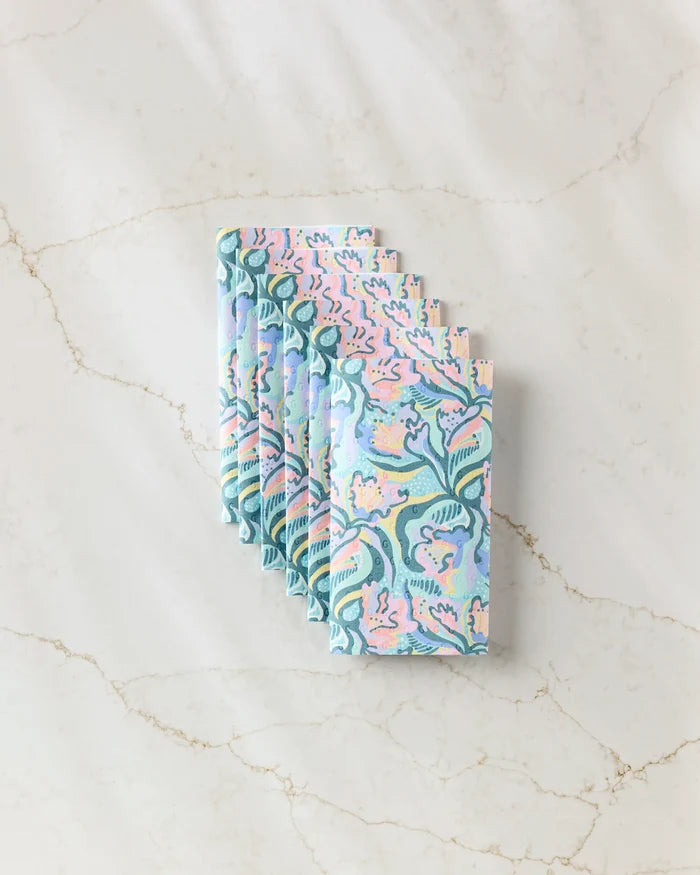 A stack of Moody Flowers napkins by Masha Volnova, made from eco-friendly paper and featuring swirling blue, pink, and green abstract foliage patterns, is arranged neatly on a marble surface.
