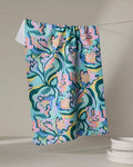 The Moody Flowers recycled kitchen towel by Masha Volnova, featuring an abstract floral pattern in pink, blue, green, and yellow, hangs on a line beside stacked white dishes on a neutral surface.