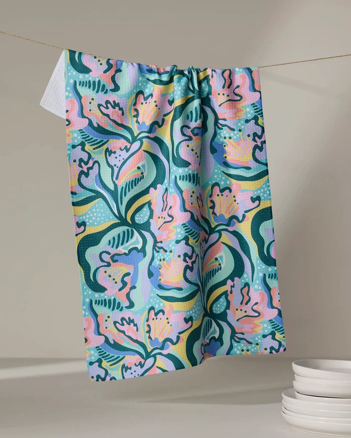 The Moody Flowers recycled kitchen towel by Masha Volnova, featuring an abstract floral pattern in pink, blue, green, and yellow, hangs on a line beside stacked white dishes on a neutral surface.
