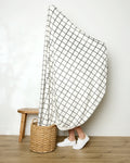 A person stands behind the large white "Moon" throw by Geometry, featuring a black grid pattern and hiding their body. Only their lower legs and white shoes are visible, with a woven basket and small wooden stool nearby.