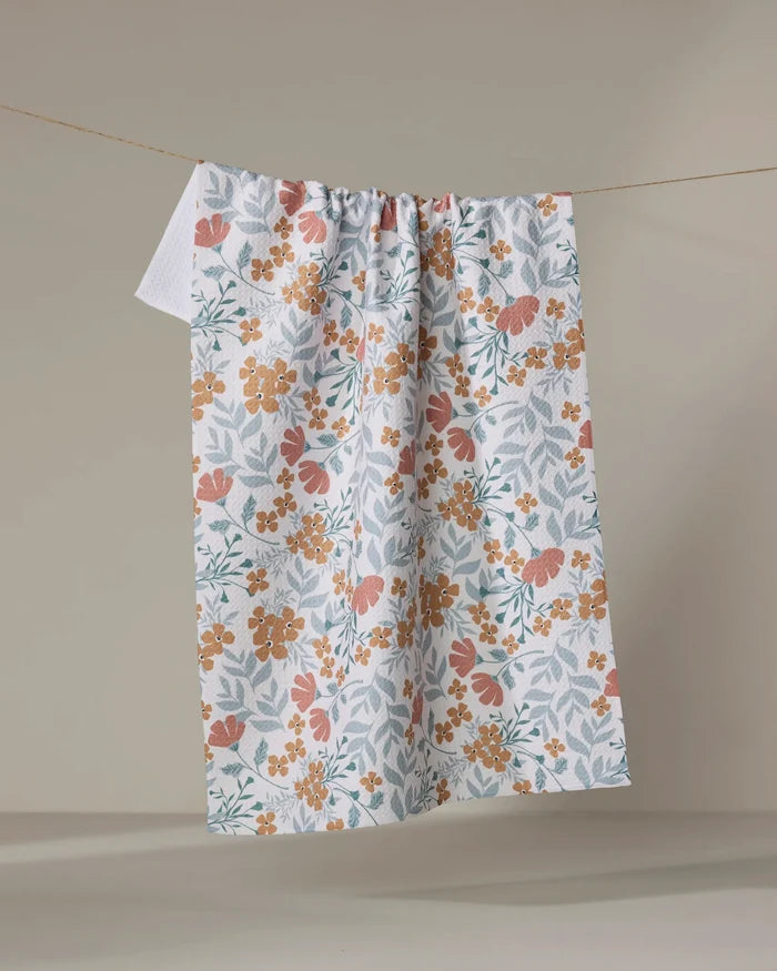 The Morning Blooms tea towel by Charlie Rowan Designs features orange and yellow florals on white, crafted from recycled materials and displayed against a neutral backdrop.