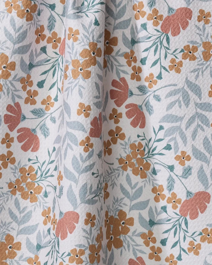 The Morning Blooms tea towel by Charlie Rowan Designs features a floral pattern with yellow-orange and pinkish-red flowers plus greenish-blue leaves on white. Made from quick-drying recycled materials, it has a textured surface and gentle folds.