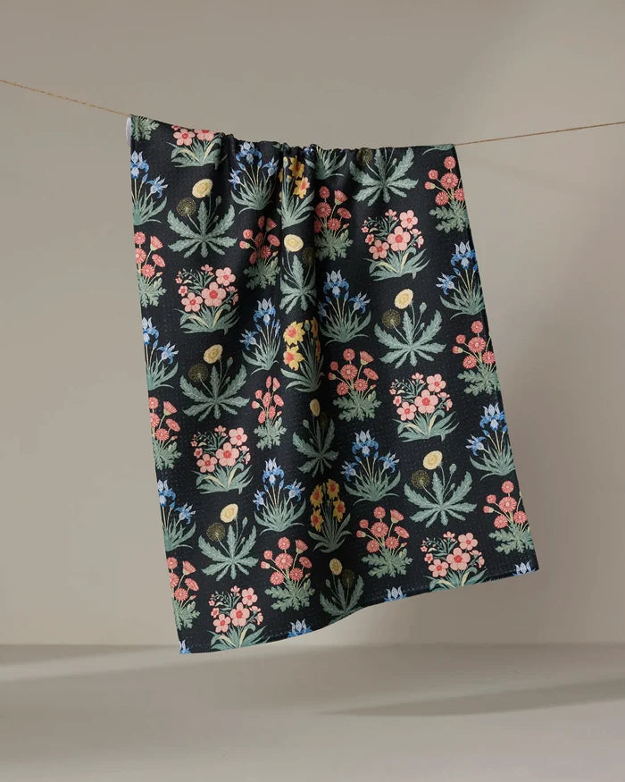 The "Morning Meadow" tea towel by Danelys Sidron, made from recycled materials, showcases vibrant floral patterns in green, pink, blue, and yellow and hangs against a neutral beige backdrop.