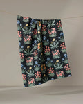 The "Morning Meadow" tea towel by Danelys Sidron, made from recycled materials, showcases vibrant floral patterns in green, pink, blue, and yellow and hangs against a neutral beige backdrop.