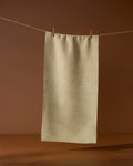 The Geometry Mosswood recycled kitchen towel in light beige hangs from a string with two wooden clothespins, set against a brown background.