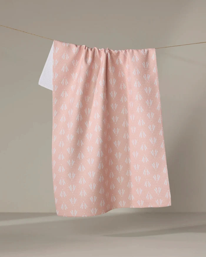 The Moth Geometric tea towel by Hufton Studio, featuring a white arrow pattern on light pink Geoweave fabric, hangs on a clothesline against a neutral beige background.