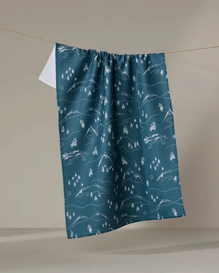 The Mountain Biking dish towel by Coit Creative features white illustrations of mountains, trees, and cyclists on a blue patterned background and hangs on a line against a plain beige backdrop.