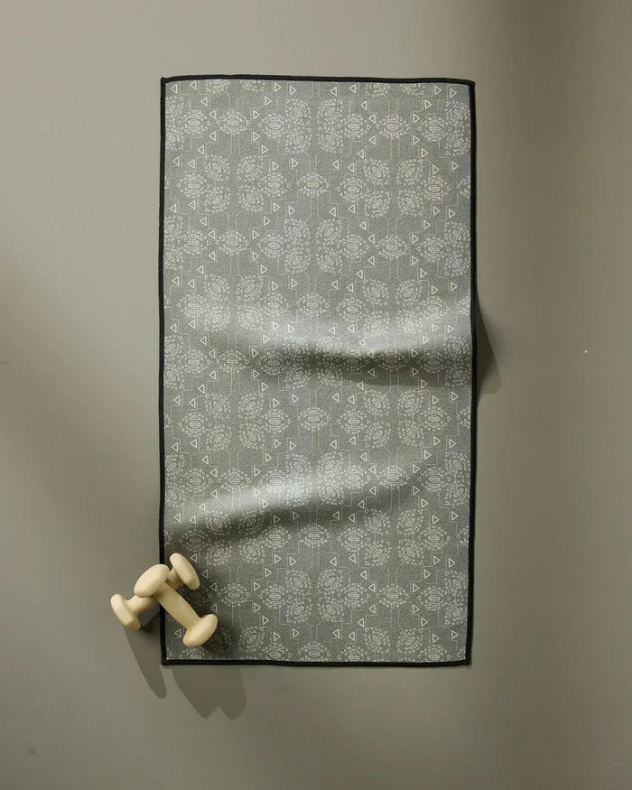 The Ash Cascade Mountain Bloom patterned grey fitness towel with a black border hangs on a wall, ultra-absorbent and fast drying, paired with small light-colored dumbbells at the bottom left corner.