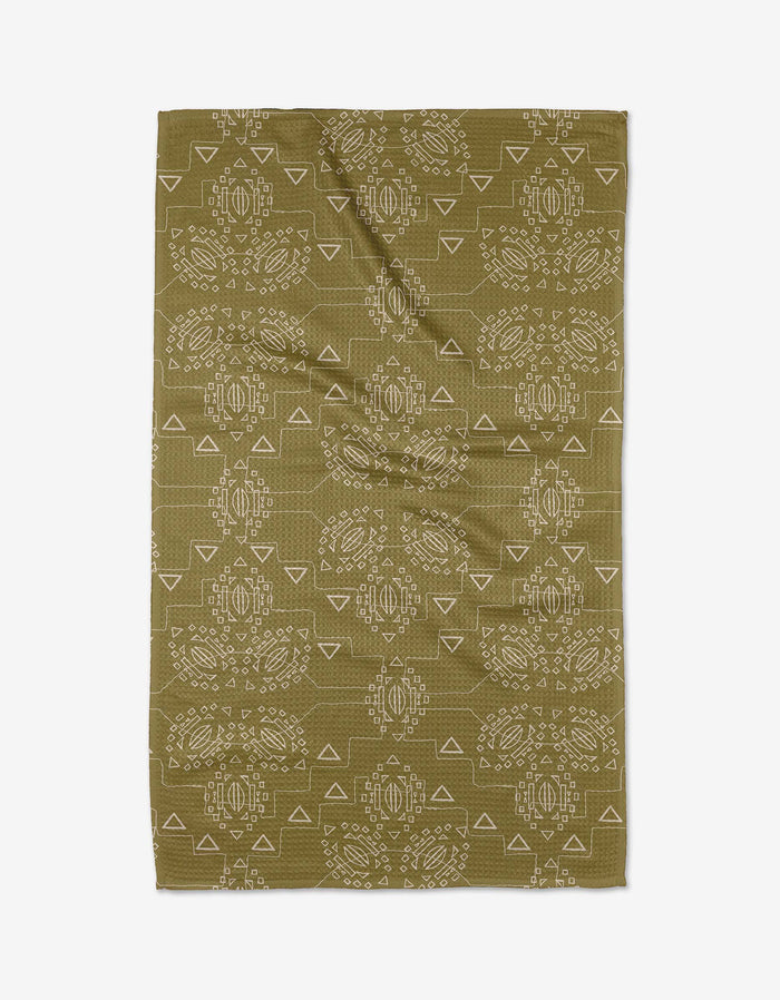 The Ash Cascade Mountain Bloom tea towel is rectangular, olive green, made from recycled materials, and features a symmetrical white geometric and tribal-inspired pattern. It is shown flat with visible creases.