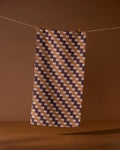 The Geometry Mountain Song bar towel features a brown, beige, and dark purple houndstooth pattern and hangs against a brown background. Made from recycled materials, it’s an eco-friendly and stylish choice.