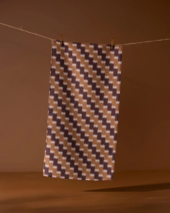 The Geometry Mountain Song bar towel features a brown, beige, and dark purple houndstooth pattern and hangs against a brown background. Made from recycled materials, it’s an eco-friendly and stylish choice.