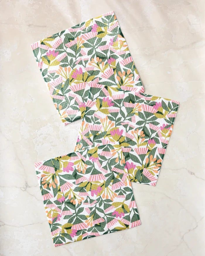 Three folded Moxie Mauve recycled dishcloths by Lisa Nelson, featuring a bright floral pattern with green leaves and pink and yellow flowers, are arranged on a light beige marble surface.