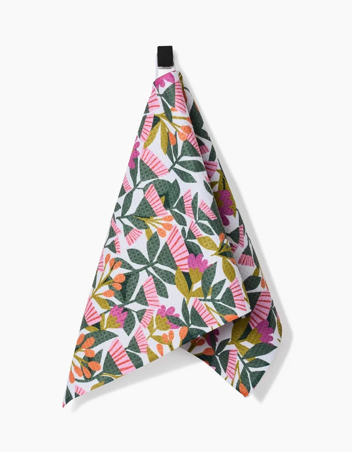 The Moxie Mauve bar towel by Lisa Nelson features a vibrant floral and leaf pattern in green, pink, orange, and purple on Geoweave fabric, shown hanging from a black loop against a white background.